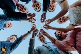 At a wedding setting in Budapest, Hungary, guests raise their glasses in celebration at a lively reception. The joyful scene captured from ground level shows friends toasting against a brilliant blue sky.